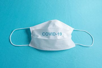 A white surgical mask with the word "COVID" is centered over a light blue background.