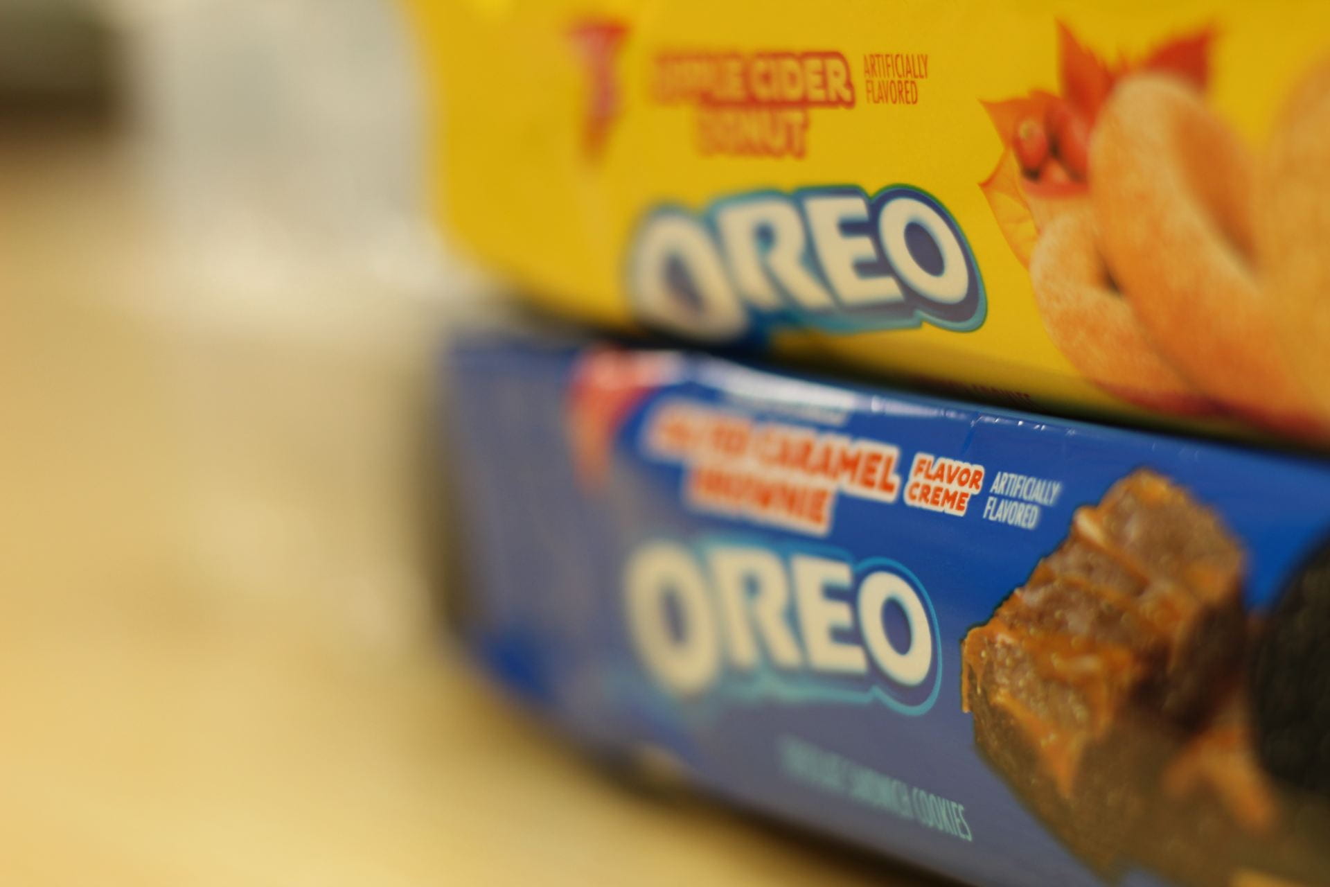 Two packages of Oreos are stacked. The bottom features a blue background with Salted Caramel Brownie Oreos. The top features a yellow background with Apple Cider Donut Oreos. The background of the photo as a whole is soft and blurred to perfection.