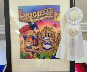 Hailey Hickerson's Battle of the Flowers band festival artwork is displayed in a black picture frame on a colorful tablecloth, garnished with a white third place ribbon.