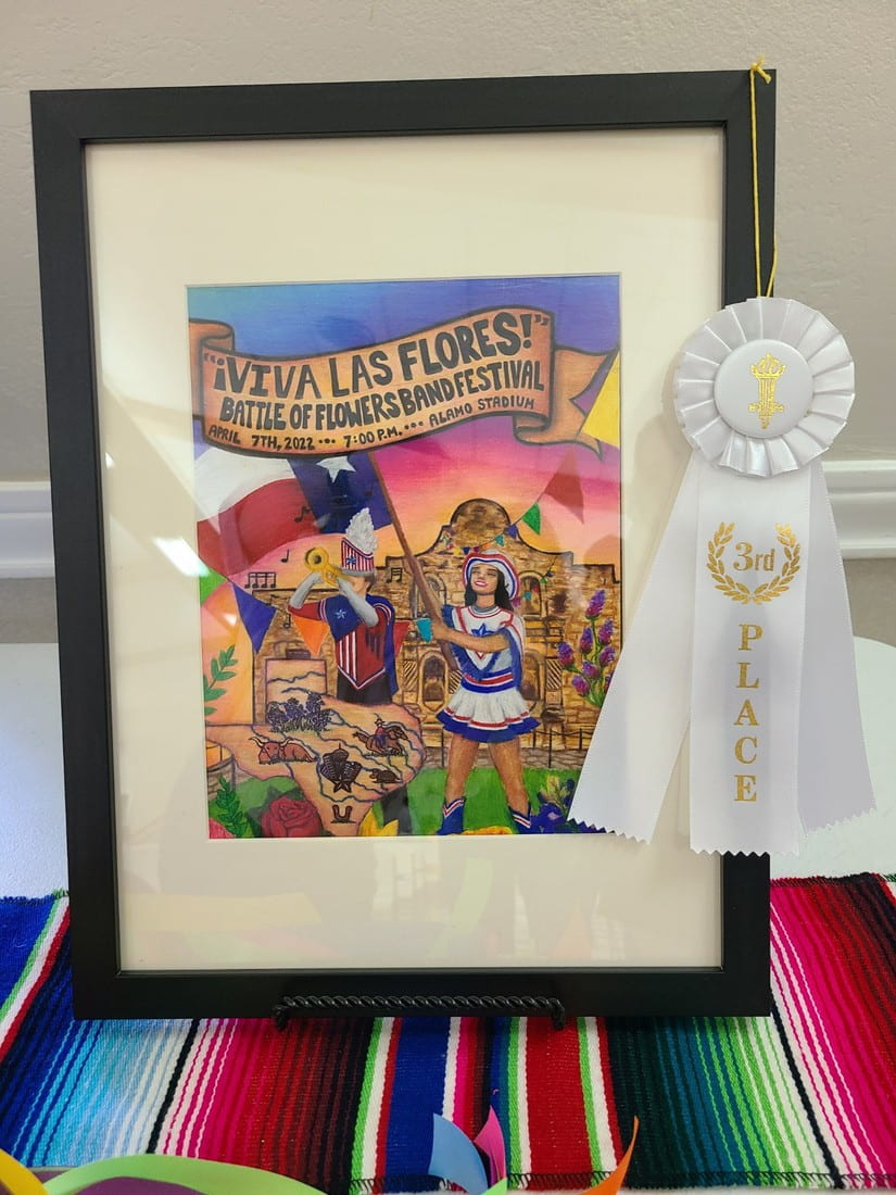 Hailey Hickerson's Battle of the Flowers band festival artwork is displayed in a black picture frame on a colorful tablecloth, garnished with a white third place ribbon.
