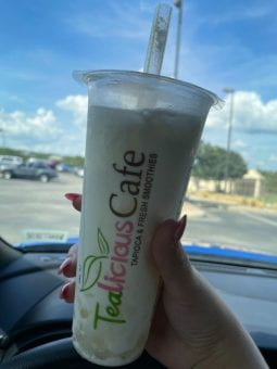 A hand is holding a boba from Tealicious cafe in a car. The logo is pink and green and the drink is lemon milk slush with lychee poppers.