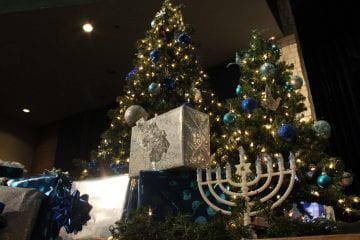 The winter decorations line the stage for the band concert. There is a lit up Christmas tree, gift boxes wrapped in festive papers, and a menorah with the Star of David in the middle.