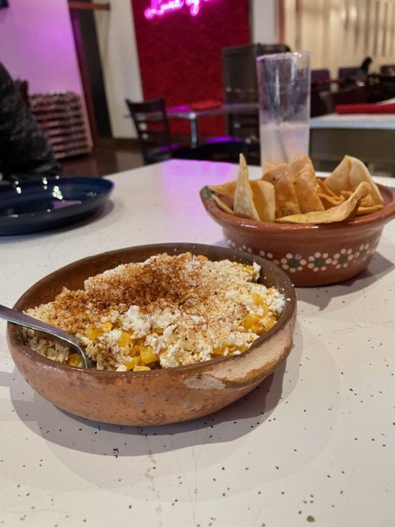 Mexican Street Corn in a stone bowl. There is a bowl of chips beside it. The corn is topped with queso fresco and chili powder.